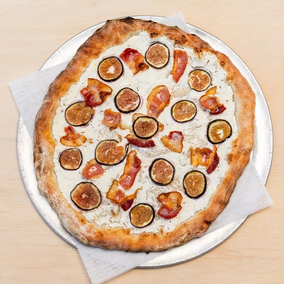 A white pizza with figs and bacon on a metal pizza pan, on a light wooden table.