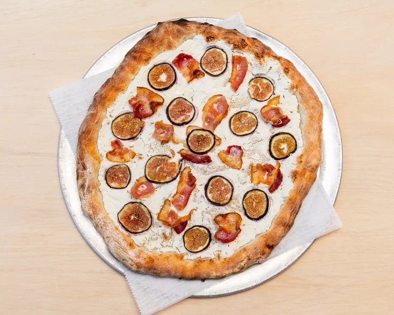 A white pizza with figs and bacon on a metal pizza pan, on a light wooden table.