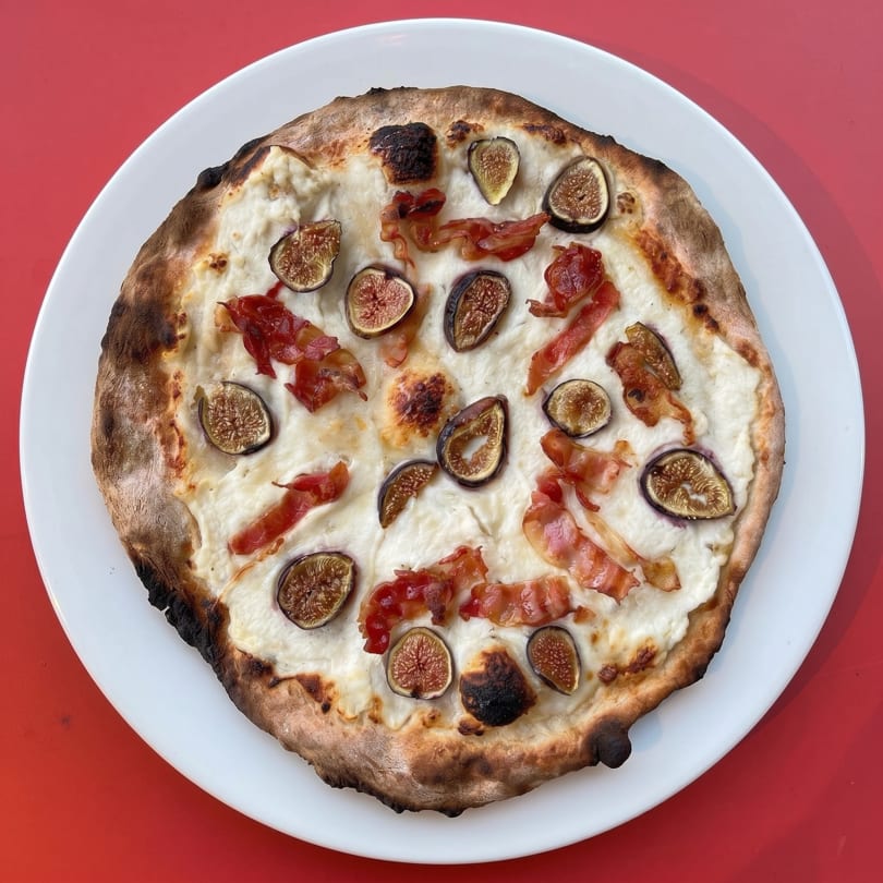 A white pizza with figs and bacon on a white plate, against a red background.
