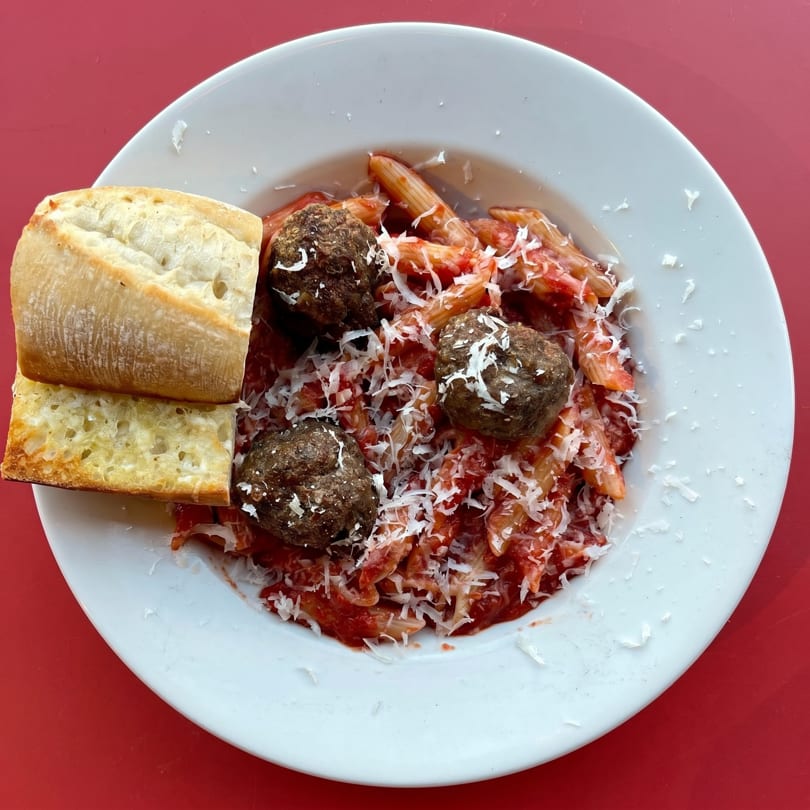 Penne pasta with meatballs, red sauce, grated cheese, and two slices of baguette on a white plate.