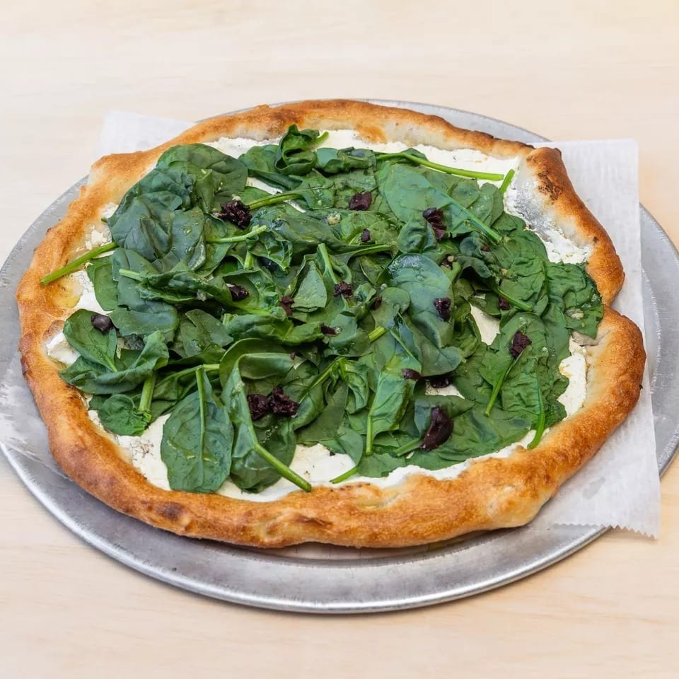 A spinach and ricotta pizza with a golden crust on a metal pan, on a light wooden surface.