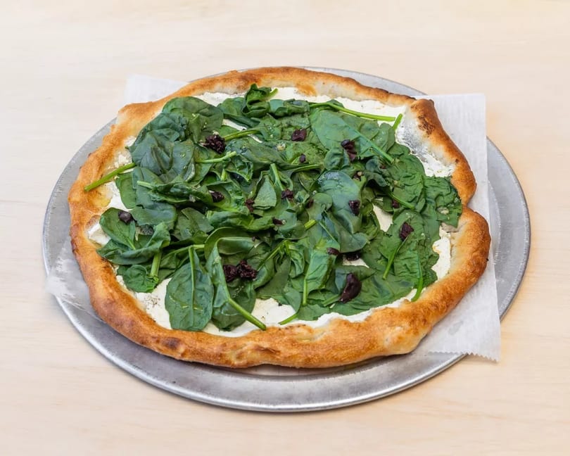A spinach and ricotta pizza with a golden crust on a metal pan, on a light wooden surface.