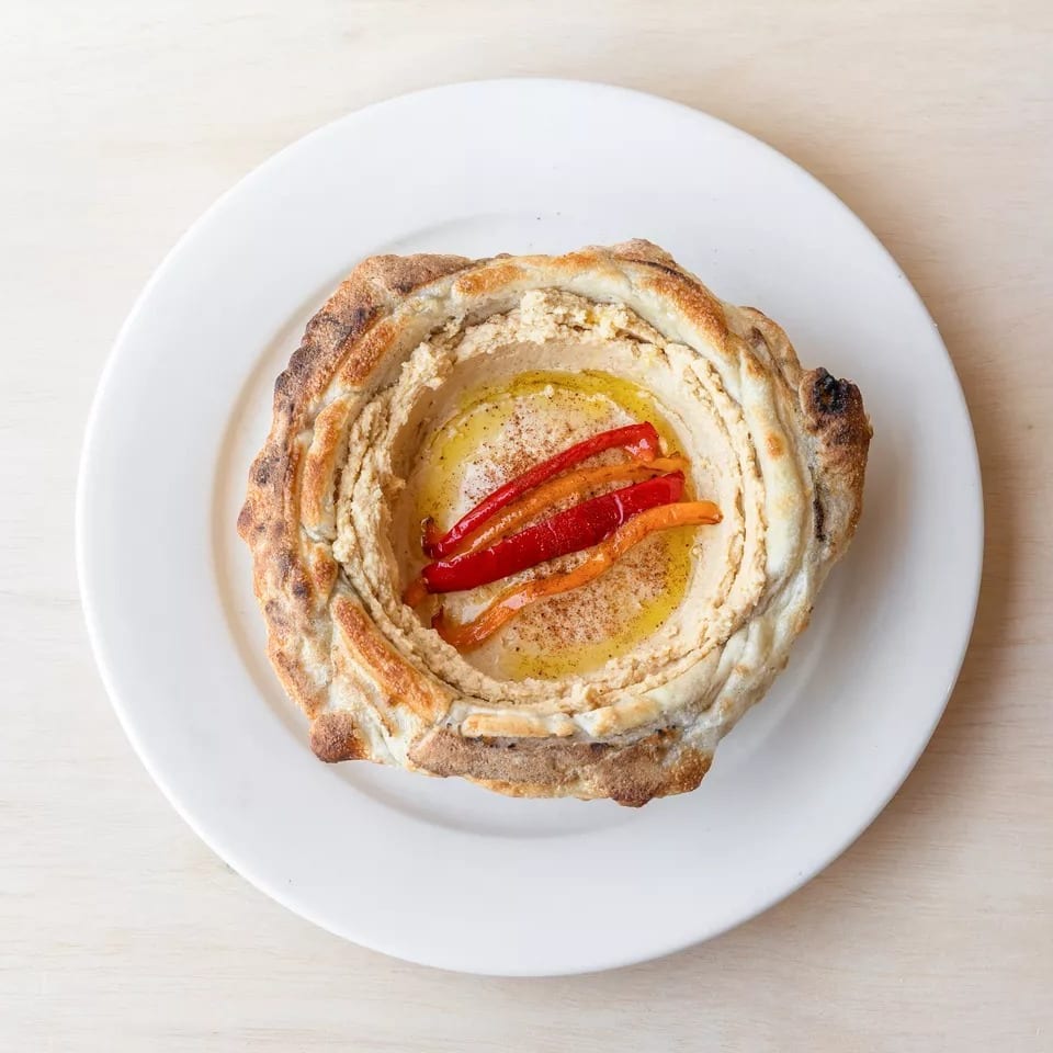 Hummus served in a baked bread bowl, topped with olive oil, paprika, and roasted red peppers.