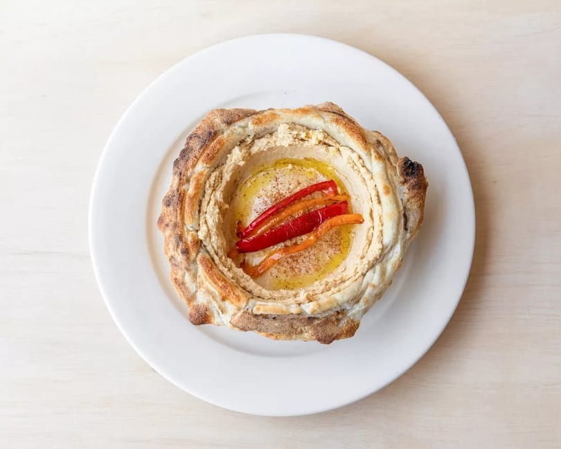 Hummus served in a baked bread bowl, topped with olive oil, paprika, and roasted red peppers.