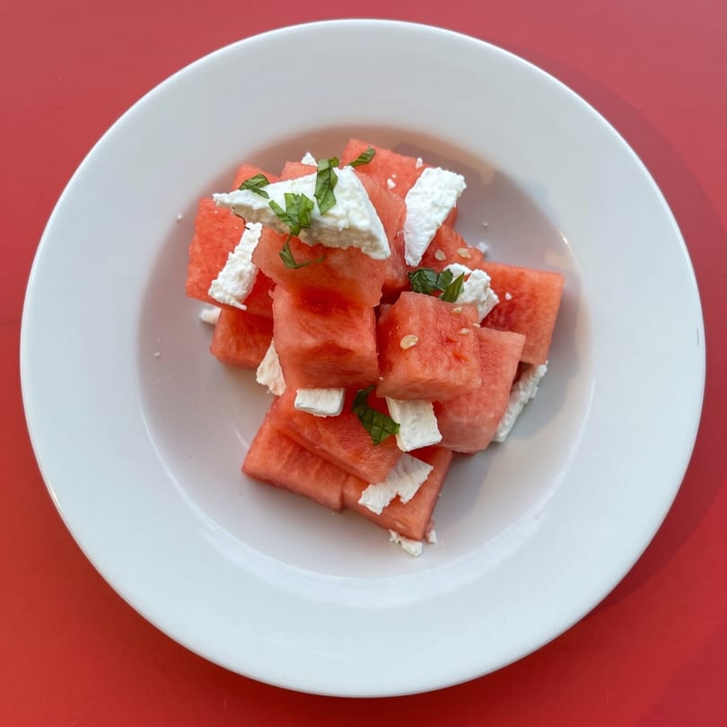 Diced watermelon with crumbled feta cheese and fresh mint leaves in a white bowl on a red surface.