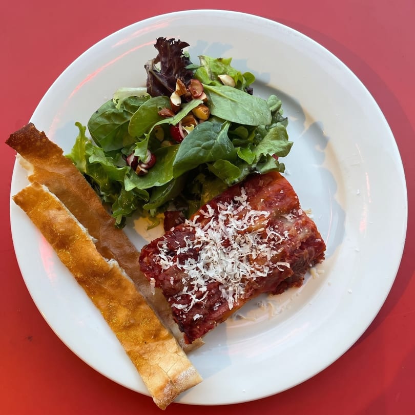 A white plate with lasagna, a green salad with nuts, and two breadsticks on a red table.