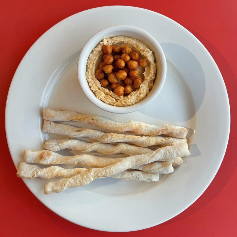 A white plate with a bowl of hummus topped with roasted chickpeas and several breadsticks.