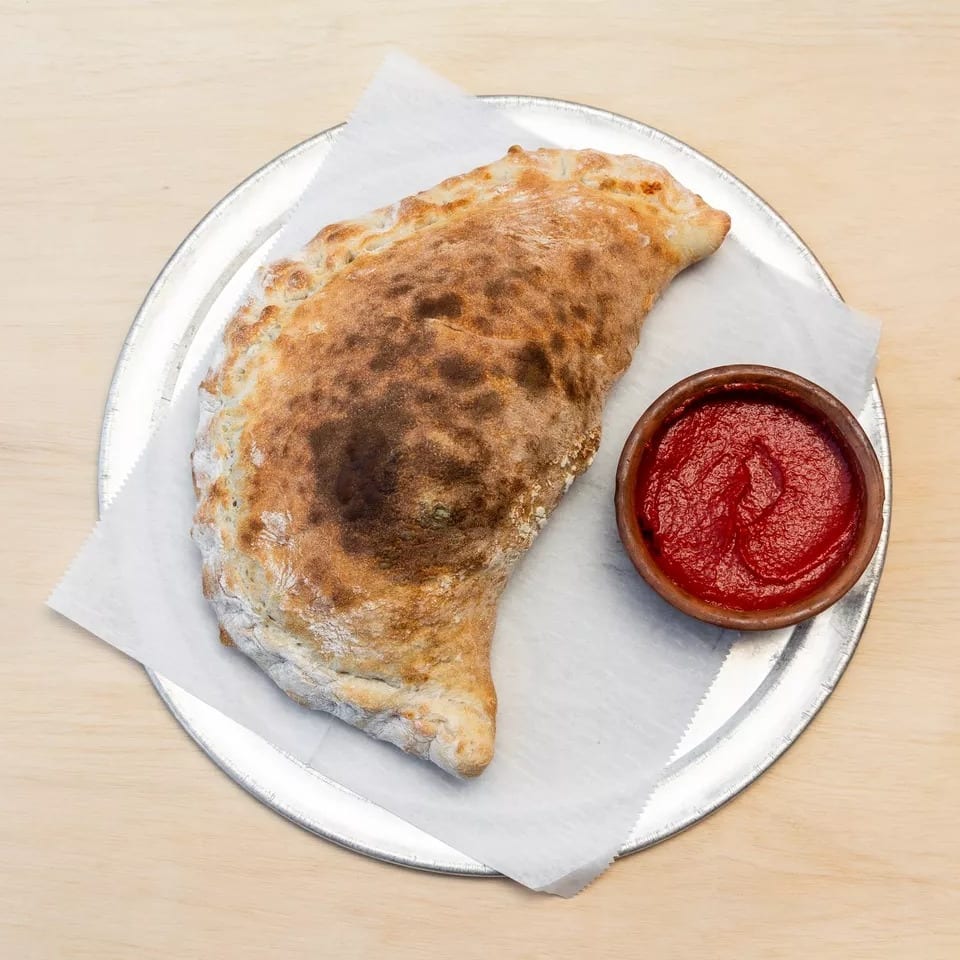 Golden-brown calzone on white parchment paper and a metal tray, with a small bowl of red sauce.