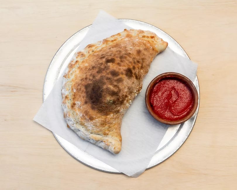 Golden-brown calzone on white parchment paper and a metal tray, with a small bowl of red sauce.