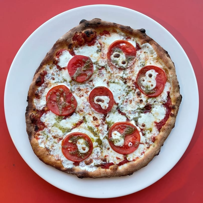 A round pizza with melted mozzarella, sliced tomatoes, and pesto on a white plate.
