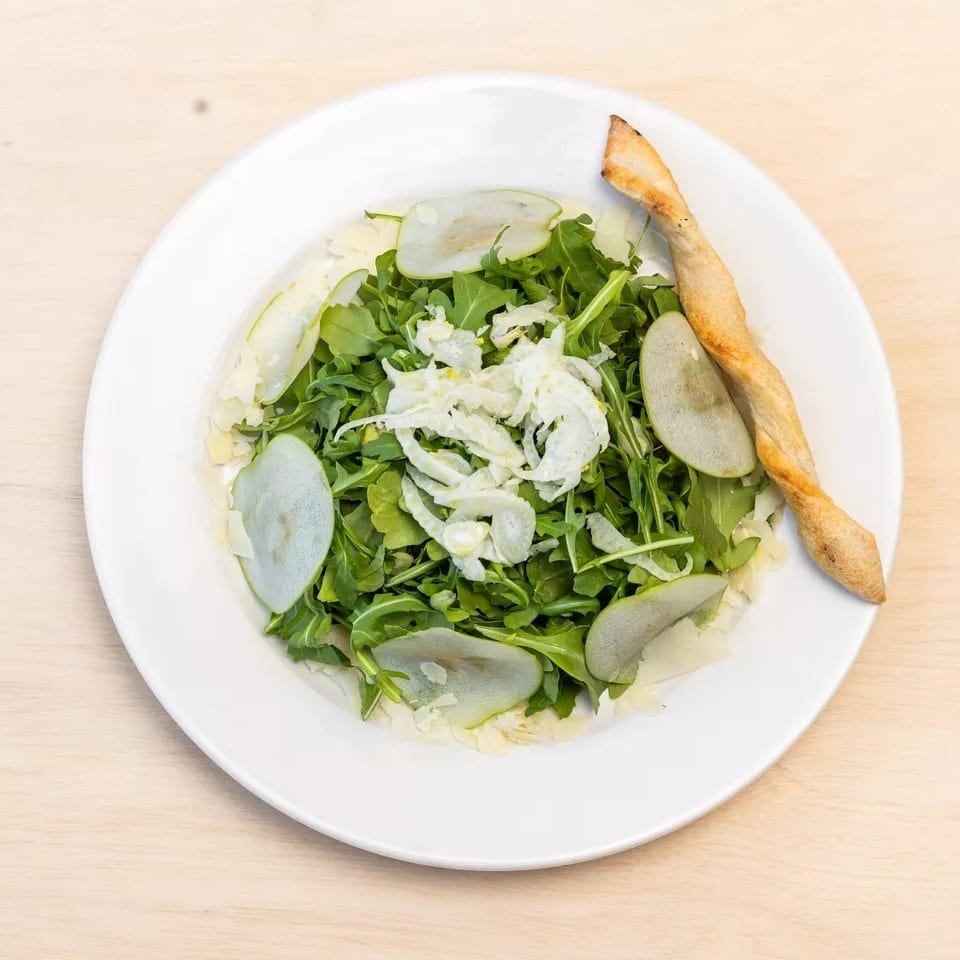 Arugula salad with sliced pears, shaved cheese, and a breadstick on a white plate.