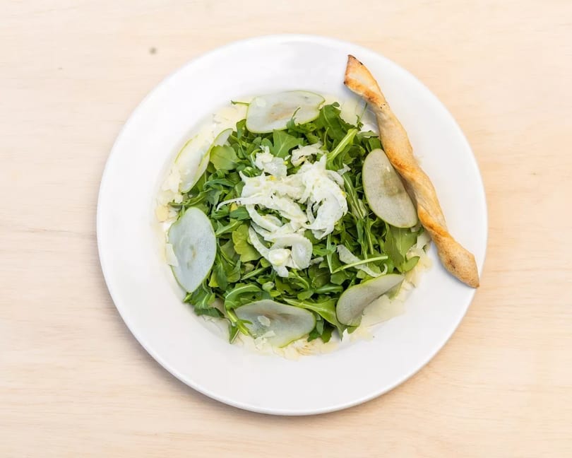 Arugula salad with sliced pears, shaved cheese, and a breadstick on a white plate.