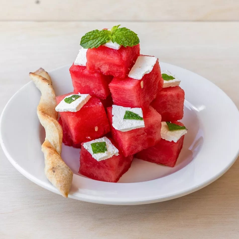 Cubed watermelon and feta cheese salad with mint and a breadstick on a white plate.