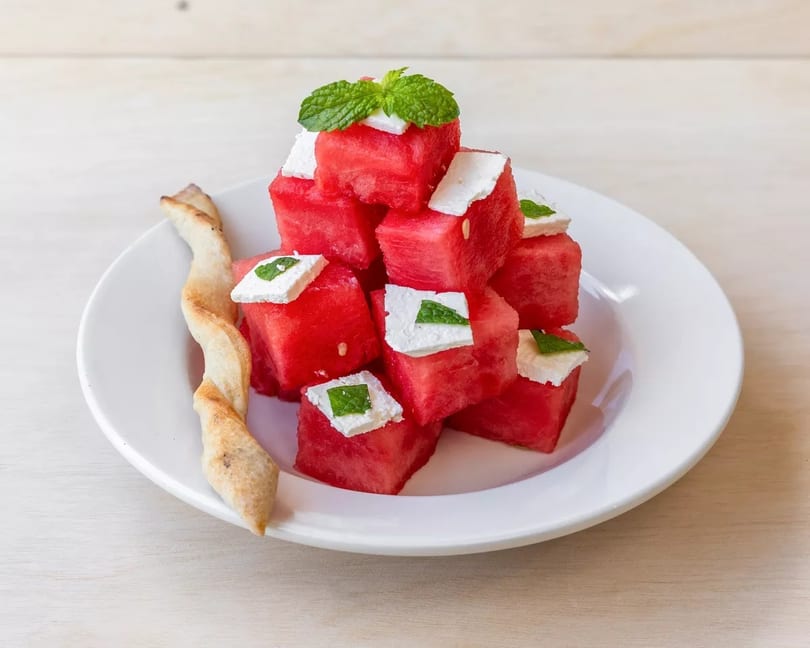 Cubed watermelon and feta cheese salad with mint and a breadstick on a white plate.