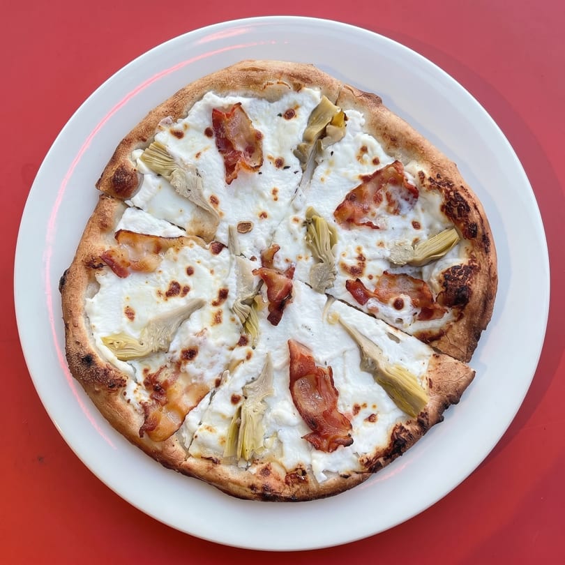 White pizza with ricotta, bacon, and artichoke hearts, sliced on a white plate against a red background.
