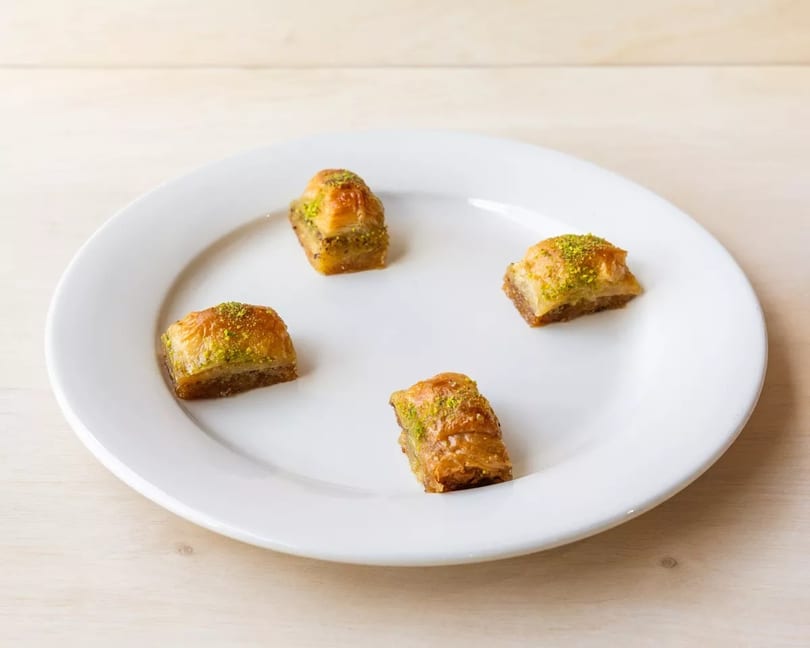 Four pieces of golden baklava with pistachio sprinkles arranged on a white plate.